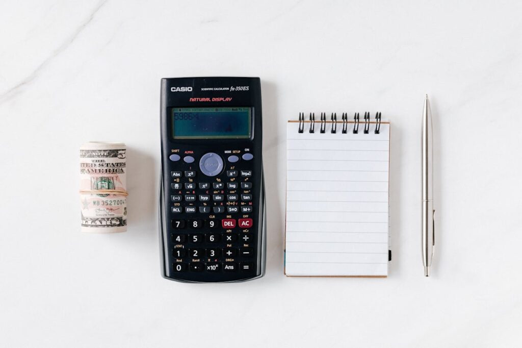 From above of roll of American dollar banknotes tied with rubber band near professional calculator and empty spring notepad with pen on marble table